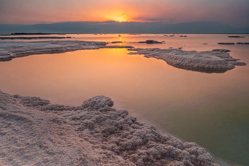 Biển Dead Sea, hay còn gọi là Biển Chết, là một trong những kỳ quan thiên nhiên độc đáo và kỳ diệu nhất trên thế giới. Nằm giữa biên giới Israel, Jordan và Palestine, Biển Dead Sea nổi bật bởi mức độ muối cực kỳ cao trong nước, khiến cho không có bất kỳ sinh vật nào có thể sống trong đó, từ đó hình thành tên gọi "Dead Sea". Nước của biển này có độ mặn gấp 10 lần so với các đại dương khác, tạo ra một môi trường kỳ lạ và đặc biệt.Điều thú vị nhất về Biển Dead Sea là khả năng nổi trên mặt nước của con người mà không cần bất kỳ nỗ lực nào. Nhờ lượng muối cao, nước biển có độ nổi cao, giúp du khách dễ dàng nằm trên mặt nước mà không bị chìm, tạo ra một trải nghiệm vô cùng độc đáo. Ngoài ra, nước biển và bùn ở Biển Dead Sea còn nổi tiếng với các đặc tính chữa bệnh và làm đẹp, nhờ vào hàm lượng khoáng chất phong phú như magie, canxi và kali. Chính vì vậy, Biển Chết đã trở thành một điểm đến nổi tiếng cho những ai muốn tận hưởng các liệu pháp chăm sóc sức khỏe và sắc đẹp. Biển Dead Sea còn có vẻ đẹp kỳ bí, với không gian rộng lớn và cát trắng mịn bao quanh. Vào ban ngày, dưới ánh mặt trời, mặt nước biển phản chiếu ánh sáng mạnh mẽ, tạo nên một cảnh tượng huyền bí và thơ mộng. Xung quanh biển là những dãy núi và sa mạc hoang sơ, làm tăng thêm vẻ hoang dại và độc đáo của khu vực này. Hình ảnh Biển Dead Sea 